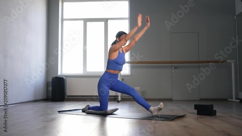 Graceful charming woman in activewear performing low lunge yoga exercise and raising both arms overhead on fitness mat, building lower-body stability, strengthening glutes and quads muscles in gym