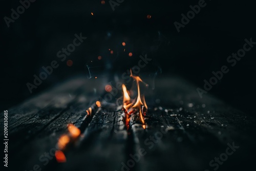 Small flames and embers burning on dark distressed wooden surface with scattered sparks and smoke against black background.