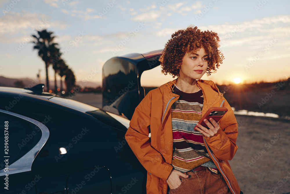Naklejka premium Woman lifestyle on road at sunset near car, curly hair, orange jacket, holding tablet, calm smile, evening light, travel portrait, parked vehicle background