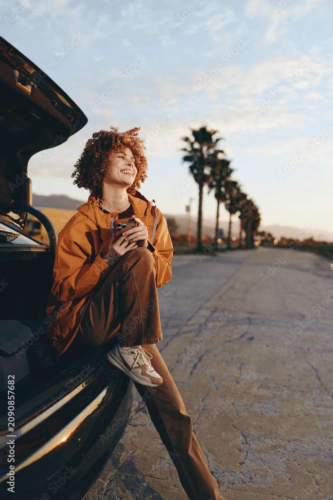 Naklejka premium Woman lifestyle outdoors near car at sunset, curly hair smiling and holding coffee, casual jacket, relaxed street mood, soft breeze, warm golden light, palm trees blurred background