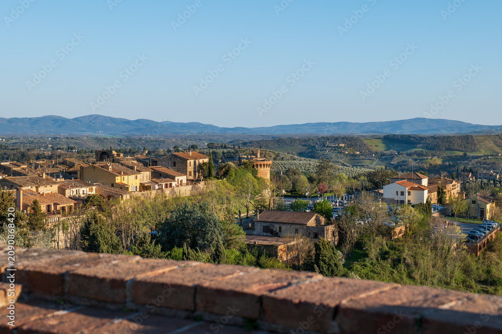 Fototapeta premium Panoramic view of a San Gimignano Tuscan village with rooftops, fields, and distant mountains under a clear blue sky. A classic rural Italian landscape.