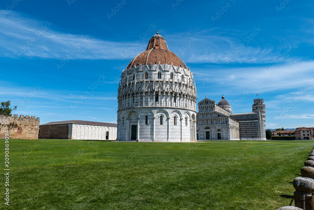 Fototapeta premium PISA ITALY APRIL 5 2026: Pisa Baptistery and the Leaning Tower of Pisa viewed from the grass under a bright blue sky. A famous Italian landmark in a classic wide-angle composition.