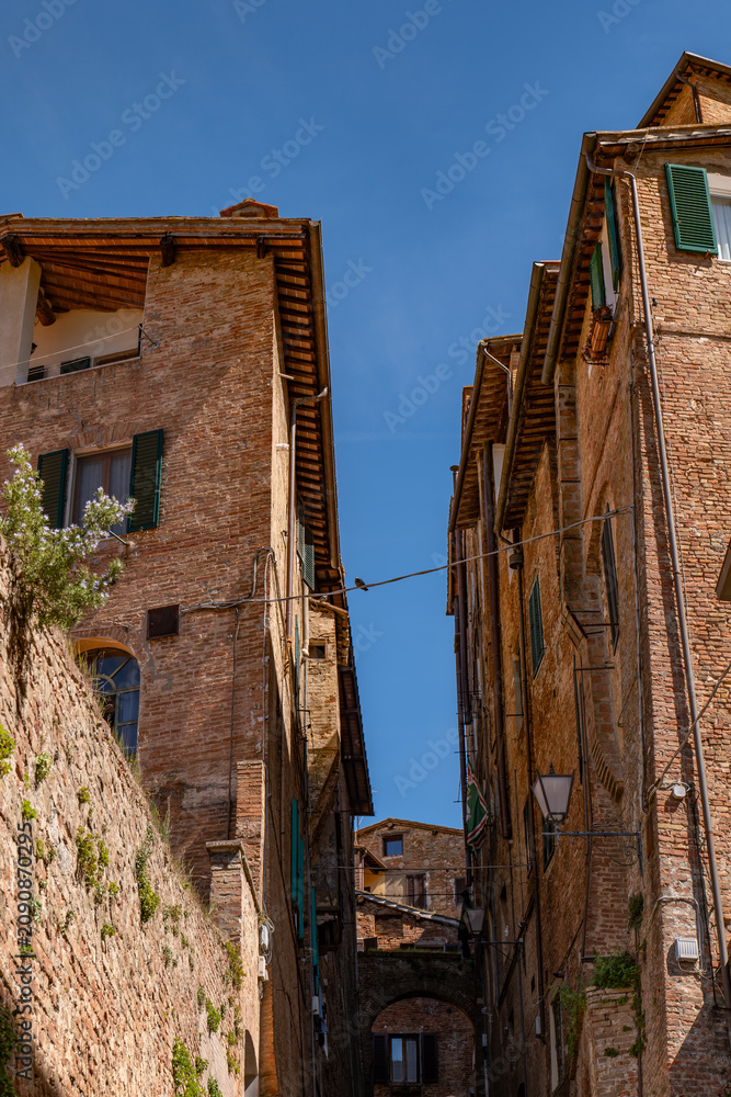 Fototapeta premium Narrow medieval street in Siena with tall brick buildings and a bright blue sky overhead. The composition emphasizes the vertical lines and old-world atmosphere.