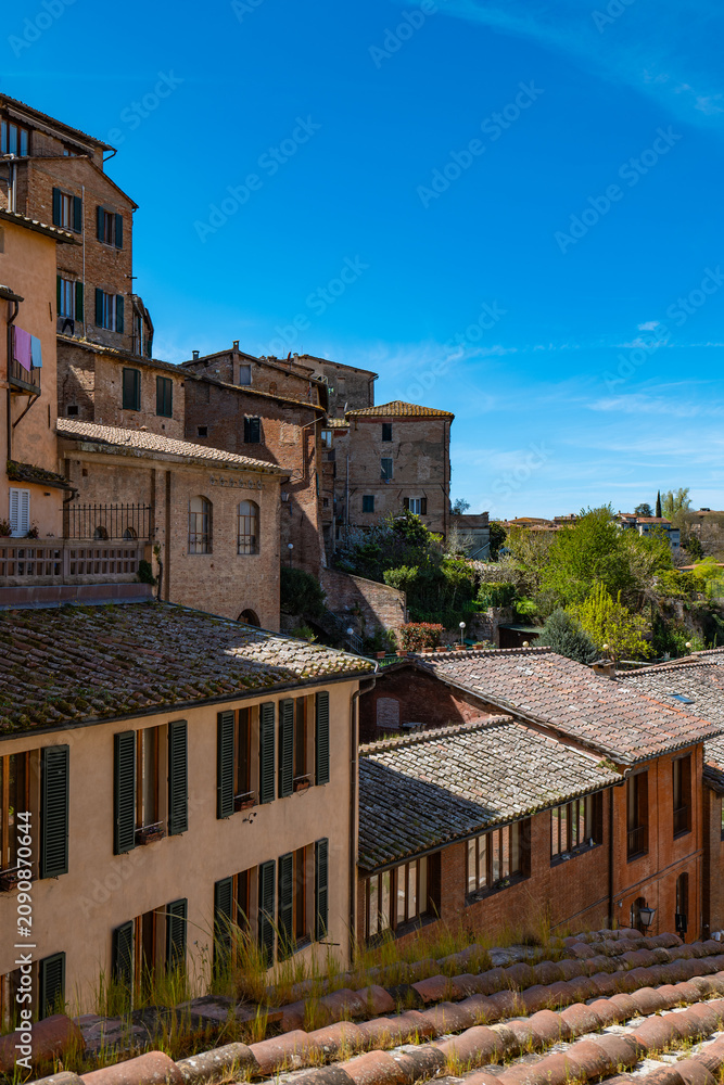 Fototapeta premium Sunlit view across Siena’s rooftops toward a prominent cathedral and historic buildings. The image captures the layered skyline of the old Tuscan city.