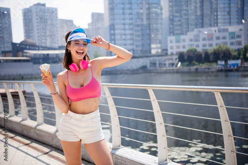 Φωτογραφία Young woman enjoying ice cream by the city waterfront in summer with pink headph