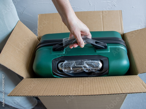 A woman's hand takes out a new green suitcase from a cardboard box.