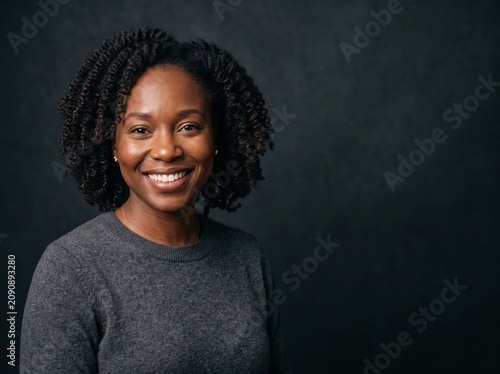 A smiling woman with dark curly hair and a grey sweater against a dark background. Her eyes are looking directly at the camera and she has a warm, inviting expression