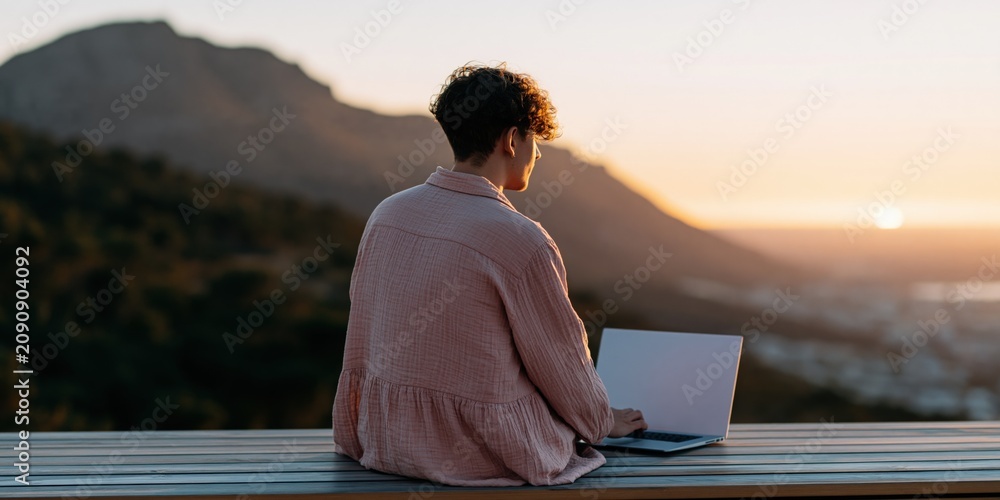 Fototapeta premium Young person working on a laptop at sunset on a mountain balcony background for remote work banner and outdoor productivity design