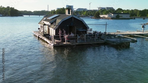 Aerial drone footage of the sauna at Soutustadion Helsinki, showcasing the sauna building, surrounding waterfront, green areas, and cityscape from above. A cinematic view of this distinctive location 