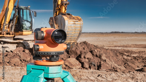 Workers use a geodetic level to measure land at a construction site. Heavy machinery operates nearby on a wide expanse of ground under a blue sky