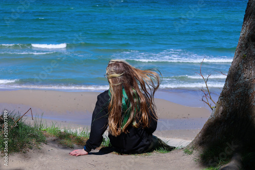 Girl sitting by a tree overlooking a sandy beach with sea waves on a windy sunny day.
