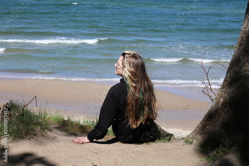 Girl sitting by a tree overlooking a sandy beach with sea waves on a windy sunny day.