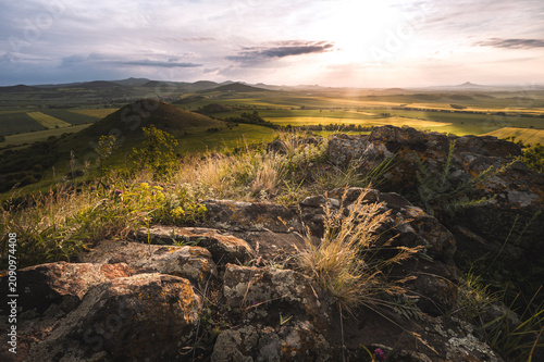 A breathtaking sunset view over the rolling hills of the Ceske Stredohori landscape, Czech Republic. Scenic volcanic peaks rise from green fields under a dramatic golden sky, captured from a rocky sum