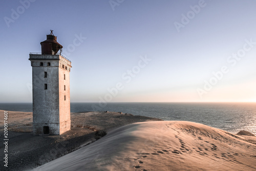 The historic Rubjerg Knude lighthouse standing on a massive sand dune by the North Sea coast in Denmark. A scenic coastal landscape captured during a peaceful sunset with clear sky.