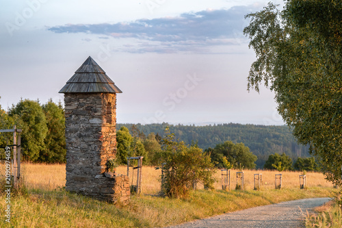 An old stone wayside shrine with a wooden roof standing next to a dirt road in the Czech countryside. A peaceful rural landscape at sunset with rolling hills and green forests in the background.