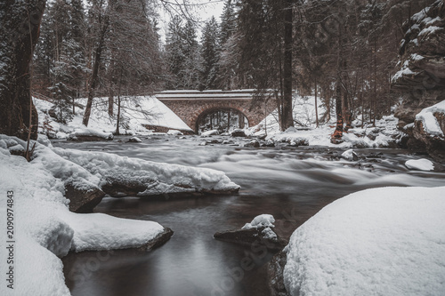 A beautiful winter landscape at Zemska Brana, Czech Republic. A wild river flows through a snow-covered pine forest under a historic stone arch bridge, captured with a long exposure effect.