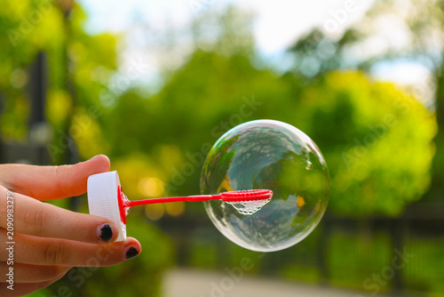Hand holding a bubble wand creating a soap bubble outdoors with a green blurred background on a sunny day.