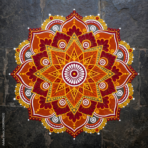 Overhead view of traditional Indian rangoli geometric mandala pattern made with colored powder in vibrant warm tones on dark floor
