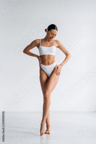 Slim female model in white bikini poses gracefully against a minimalist background, showcasing an ideal body shape and emphasizing fitness and health aesthetics