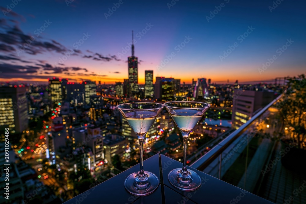Fototapeta premium Two elegant cocktail glasses on a rooftop balcony at sunset with a glowing city skyline backdrop for luxury hospitality and nightlife branding