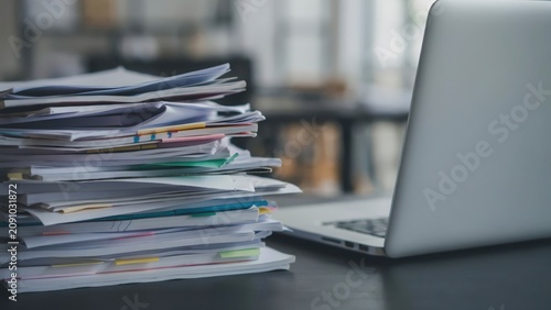 Stack of paper documents and a laptop computer on a desk in a modern office room