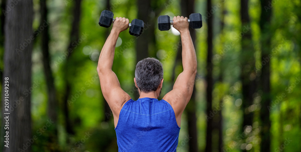 Obraz premium Strong man do sport exercise. Outdoor workout. Sport athlete man with dumbbell. Healthy lifestyle. Biceps muscle. Strong triceps. Man exercising with dumbbell. Man athlete lifting dumbbell, back view