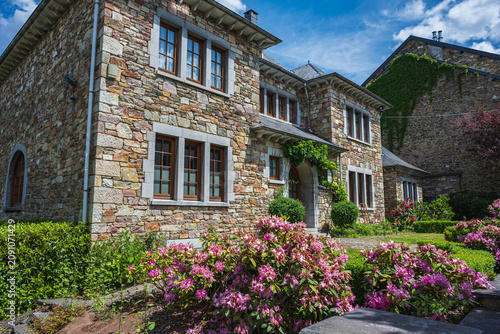 Beautiful traditional stone house in Houffalize, Belgium, with a blooming garden full of pink rhododendron flowers under a blue summer sky.