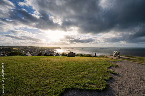 Panoramic sunset view over Gold Beach and Arromanches-les-Bains from the cliff in Normandy, France, featuring World War II remnants and cloudy sky.
