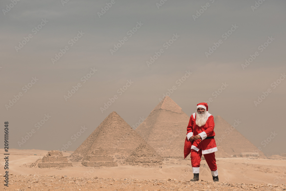Fototapeta premium Iconic pyramid backdrop frames a surreal and festive Santa Claus in full red costume holding a gift bag on the sandy Giza plateau under a hazy
