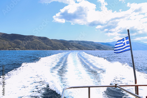 Boat wake trailing across bright blue sea beside Greek flag, mountain coastline, open sky, white clouds, shimmering water, stern railing and travel scenery, with boat, wake, sea, flag detail