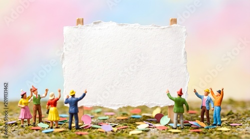 Miniature figures cheering around white torn paper signboard with colorful confetti announcement concept