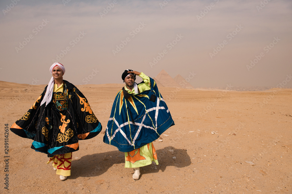Fototapeta premium Two Egyptian whirling dancers wearing vivid embroidered traditional costumes strike theatrical poses on the Giza desert plateau with the iconic