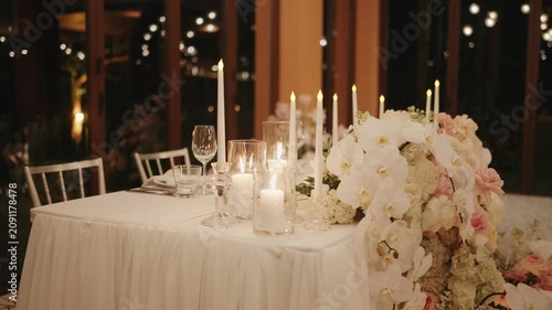A beautifully decorated wedding sweetheart table covered in flowing white fabric, adorned with lush pink and white floral garlands and glowing candles, perfect for romantic celebrations.