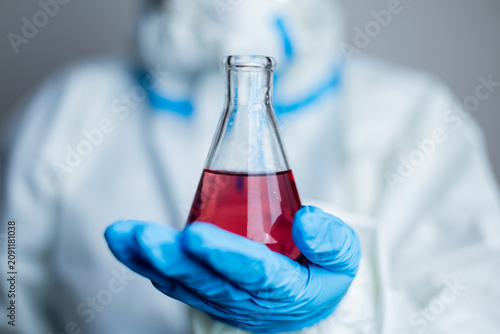 Obraz na płótnie Scientist holding a laboratory flask containing red liquid while wearing protective equipment, representing scientific research, chemical analysis, and laboratory innovation