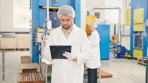 Handsome Caucasian male worker wearing lab coat and protective cap holding clipboard and pen. Looking at large industrial machinery and filling out production data. Colleges working at the background.