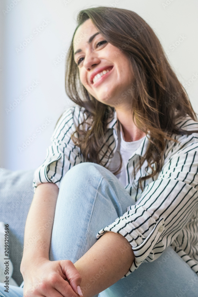 Fototapeta premium Smiling young woman sitting casually at home, relaxed and happy in a striped shirt and jeans, enjoying a peaceful moment