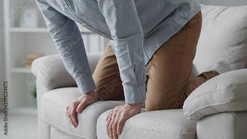Middle aged man getting up from beige sofa at home feeling back pain while kneeling on cushion seat.