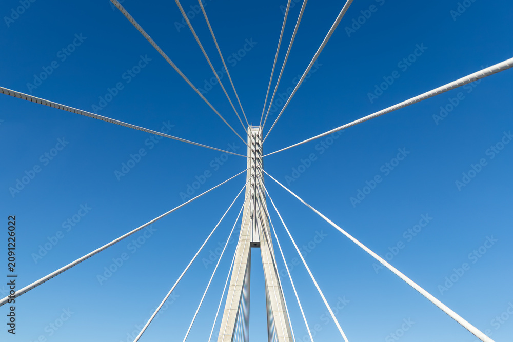 Fototapeta premium Cable bridge pylon rises into clear blue sky with steel supports forming symmetrical geometry. Minimal industrial view highlights engineering infrastructure tension modern design, and Germany travel
