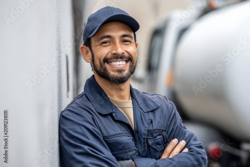 Portrait of a smiling propane technician, hispanic guy, in uniform standing beside a propane tank or truck, neutral and uncluttered background, warm approachable tone.