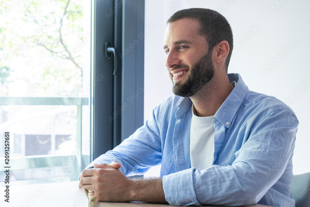 Fototapeta premium Smiling young man in a blue shirt sitting by a window, relaxed and thoughtful in a bright modern home or office setting