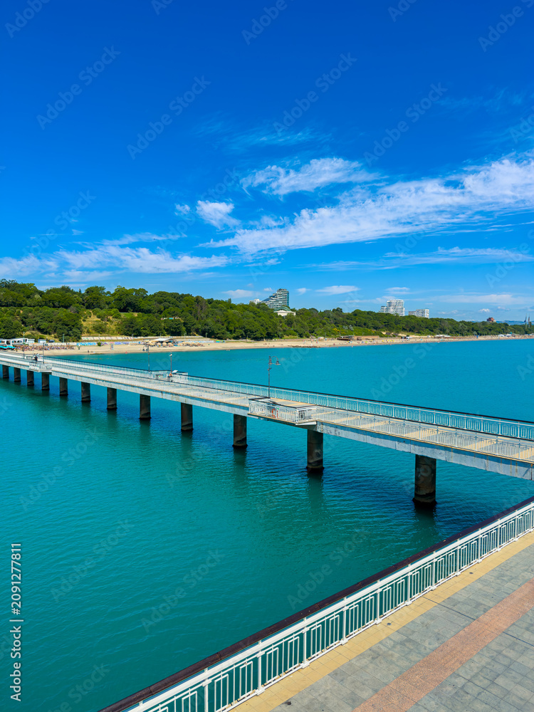 Fototapeta premium Pier extends into blue water under clear sky near green shore with buildings in the distance on a sunny day