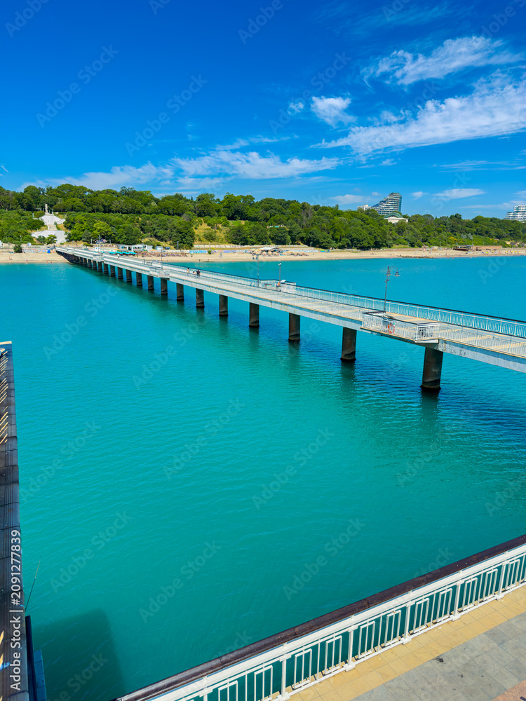 Fototapeta premium Pier extends into blue water with clear sky and green hillside in the background during bright daylight in an outdoor location