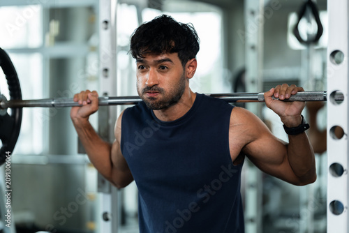 Young Asian man holds barbell across shoulders at squat rack in modern gym, pausing to catch breath during strength workout, fitness recovery, healthy lifestyle, wellness motivation concept.