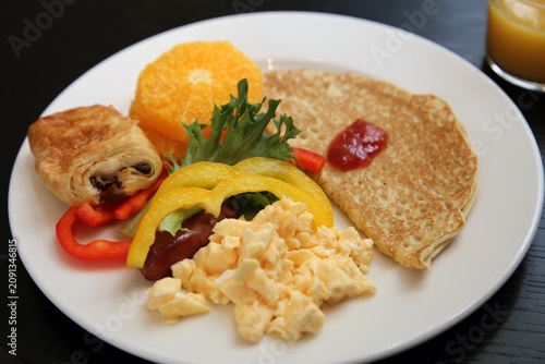 Delicious Nordic hotel breakfast including scrambled eggs, veggies, crepes with berry jam, fresh fruit, chocolate croissant and orange juice. Served on a black hotel table.