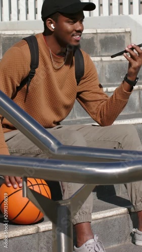 Vertical video: Raising phone, student wearing cap calling friend on steps, resting on basketball
