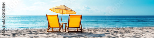 Wide view of two empty orange lounge chairs under a yellow parasol on a sandy shore facing the calm blue ocean