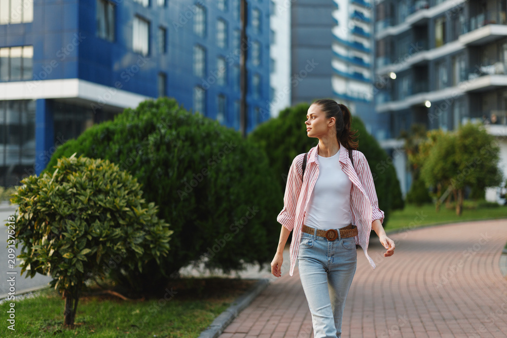 Fototapeta premium Young woman strolls along a sunny city street in jeans and a pink plaid shirt, radiating confident student energy as she walks past glass towers and green spaces.