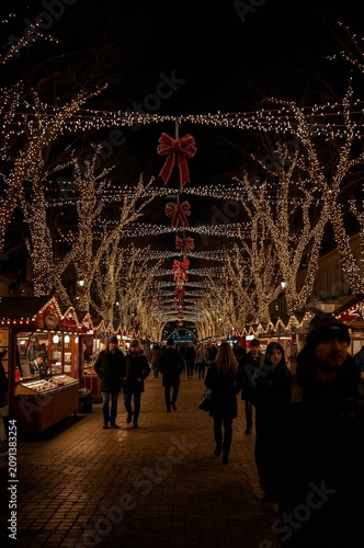 People Walk Through a Market Street With Lights and Stalls During Evening Tim...