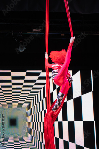 Canvastavla A female aerial gymnast wearing a red and checkered costume with a bright red wig executes a flexible vertical split while suspended on hanging red silks during a stage performance