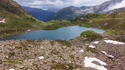 Aerial view of a turquoise alpine lake nestled in rocky tundra terrain with patches of snow, green meadows, and a wide mountain valley opening into the Pyrenees, Andorra